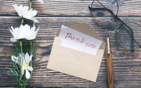 A "thank you" note card on a desk with a small bouquet of daisies, a pair of glasses, and a pen.